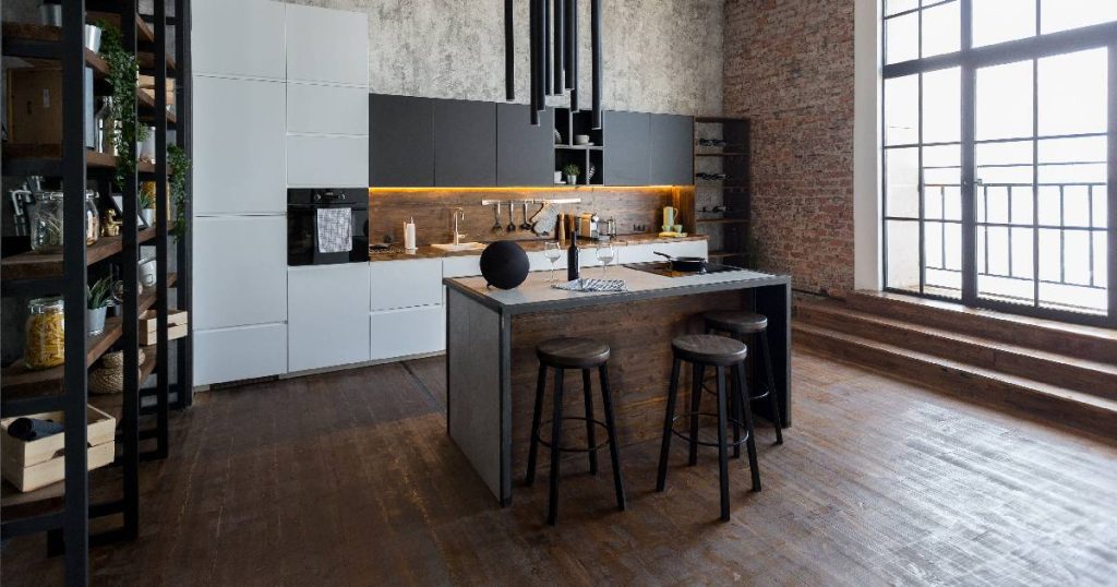 Een industriële keuken met een strakke, moderne en minimalistische look. De keuken beschikt over een grote kastenwand met spoelbak, een grote, open zwarte stellingkast en een robuust bakstenen kookeiland met kookplaat en toog. De houten vloer gaat over in lage trapjes die uitgeven op een terras achter een groot fabrieksraam. De betonnen wand achter de hoge kasten en de stoere hanglampen dragen bij aan het industriële karakter.