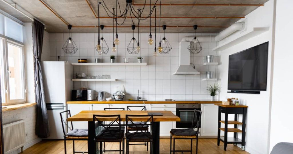 Een keuken in een appartement in een eenvoudige, industriële stijl. De keuken beschikt over een witte tegelwand, eenvoudige apparaten en robuuste hanglampen met zichtbare koperen leidingen. Een houten tafel met stoelen met zwarte metalen frames maakt het plaatje compleet.