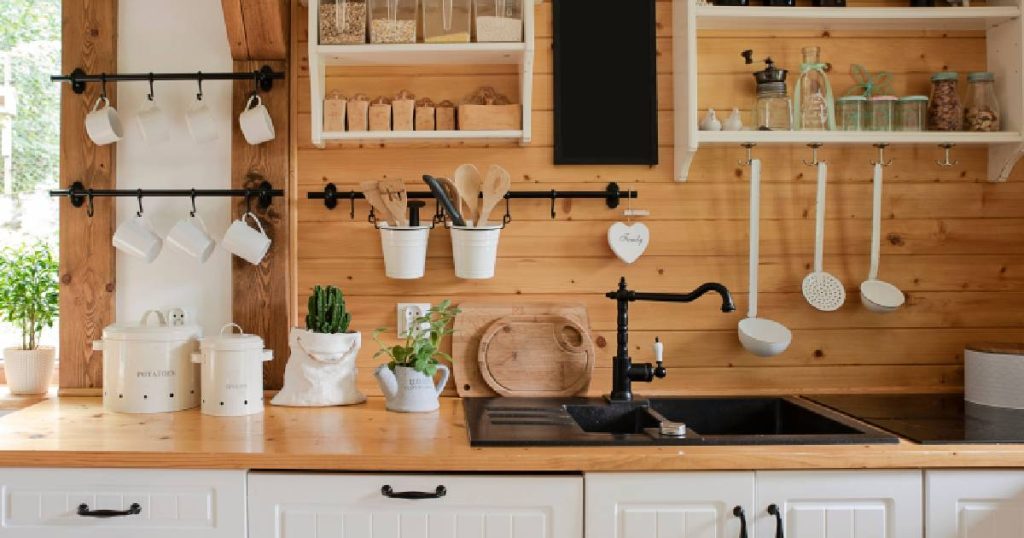 Een gezellige landelijke keuken met houten achterwand, witte hangkastjes waar kopjes en keukengerei aan hangt, en een leuke zwarte spoelbak met speelse zwarte kraan. 