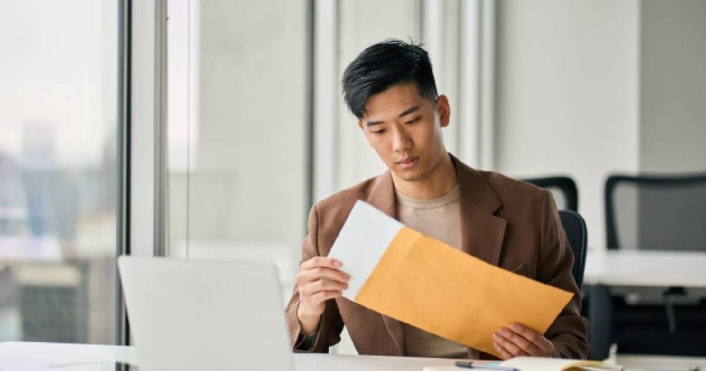 Een jonge man met zwart haar, bruin t-shirt en donkerbruin vest opent een envelop waarin het inschrijvingsbewijs of kentekenbewijs van zijn nieuwe auto zit.