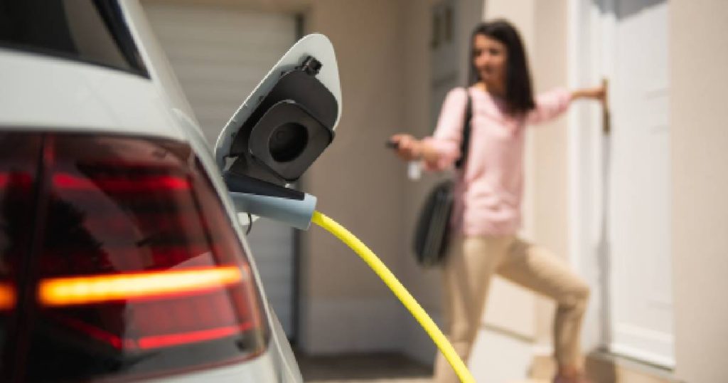 Een witte elektrische auto wordt opgeladen via een gele laadkabel van een laadpaal voor thuis - die fiscaal aftrekbaar is. Op de achtergrond opent een dame in roze hemd en beige broek de deur van haar woning terwijl ze de wagen vergrendelt.