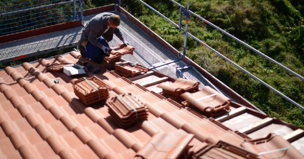 Een dakwerker legt vlakke keramische dakpannen op het dak van een woning. Hij gebruikt een steiger om het dak veilig te bereiken.