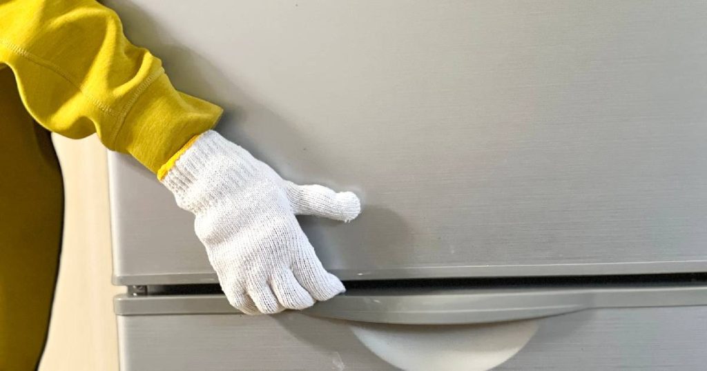 Een persoon in gele trui met witte handschoenen tilt een oude koelkast op tijdens het demonteren van de keuken.