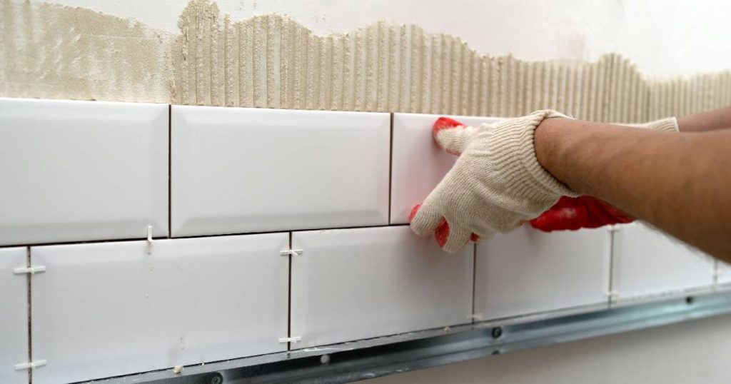 Een man met witte handschoenen is witte tegels aan het plaatsen als spatwand in de keuken.