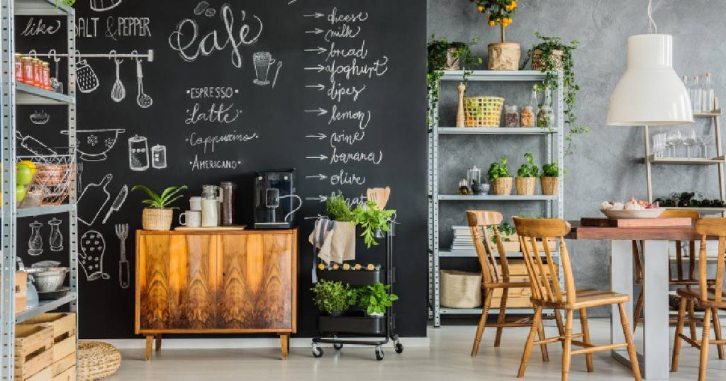 Een industriële keuken met betonlook muren, een gietvloer, een houten keukenkast en tafel met houten stoelen en verschillende metalen rekken. Een grote zwarte muur met bordverf werd gebruikt om ingrediënten en tekeningen tentoon te stellen.