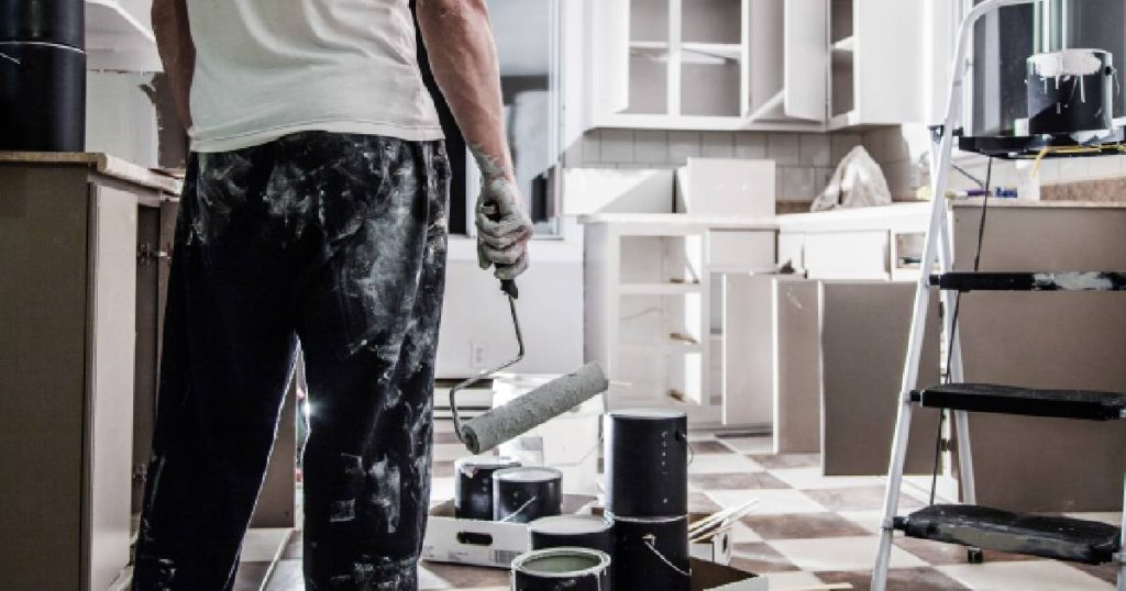 Een keukenrenovatie expert staat in een net geschilderde keuken. Hij heeft een verfrol vast en draagt een zwarte broek met witte verfvlekken en een wit t-shirt. Naast de vakman staat een trapladder.