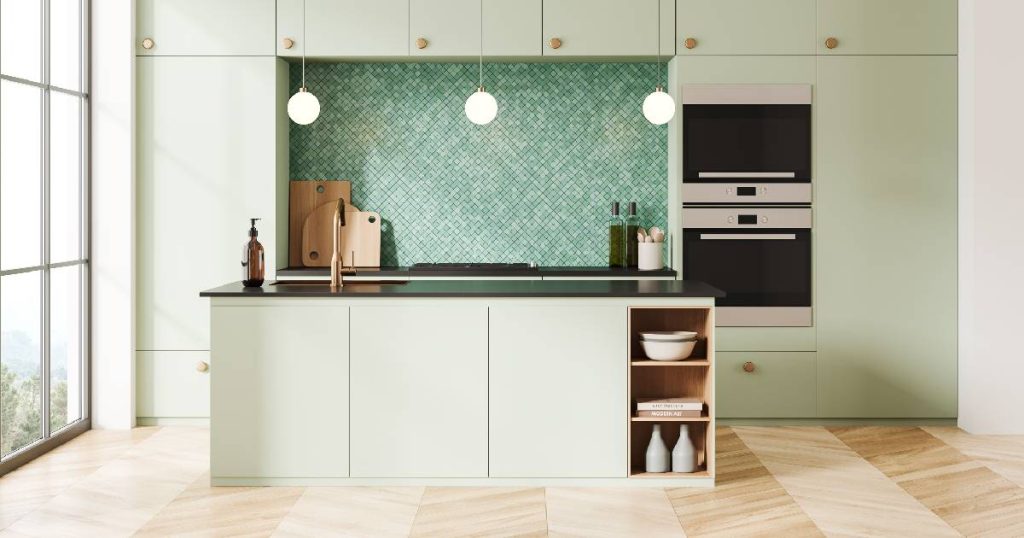 Een relatief kleine tot gemiddeld grote keuken met de ideale afmetingen voor een comfortabel eiland met spoelzone en opbergruimte. De keuken, met mintgroene keukenkasten, ingebouwde apparatuur en houten vloer, wordt door een groot raam voorzien van veel natuurlijk licht. De spatwand in kleine groene mozaïektegeltjes geeft de keuken een authentieke toets.