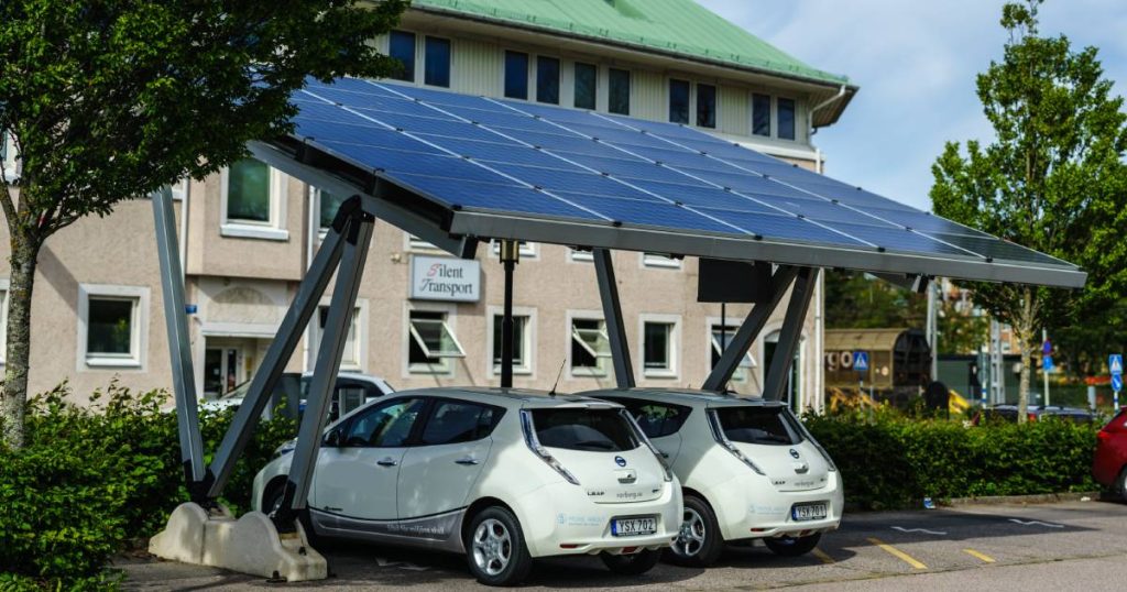 Een carport met zonnepanelen waaronder twee witte elektrische wagens opladen aan een laadpaal.