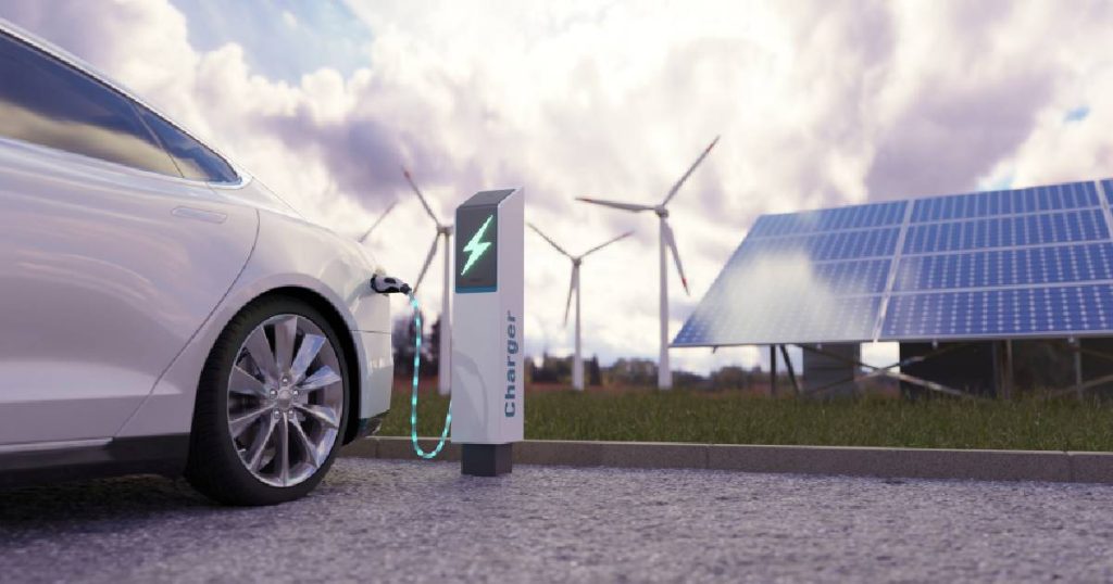 Een witte elektrische wagen en een witte laadpaal op een parkeerterrein met kiezelvloer, op de achtergrond grote blauwe zonnepanelen en witte windmolens tegen een zonnige maar bewolkte blauwe hemel.