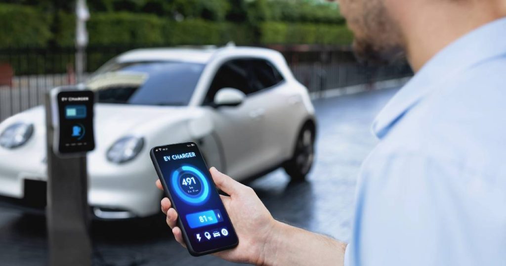 Een man met een gsm in de hand. Op het scherm van zijn smartphone volgt hij het laadproces op via een handige applicatie. Op de achtergrond de laadpaal en een witte elektrische auto.