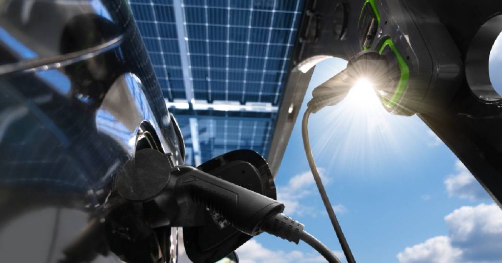 Een elektrische auto naast een laadpaal onder een carport met zonnepanelen bij de bestuurder thuis. Boven de carport een heldere blauwe hemel met kleine wolkjes en de stralende zon.