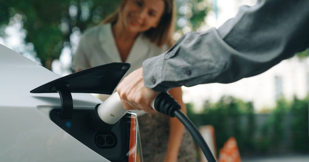 Een arm in grijs hemd steekt de laadkabel van een laadpaal voor thuis met gepast vermogen in een lichtgrijze elektrische wagen. Op de achtergrond een dame in wit hemd.