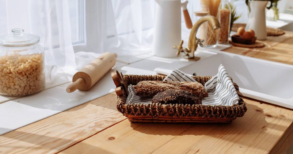 Een landelijke keuken met een werkblad uit eikenhout en rustieke accessoires: een porseleinen spoelbak met koperen kraanwerk, een rieten mandje met ambachtelijk gebakken brood, een houten roller, glazen pot en porseleinen kruiken.