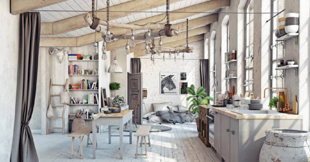 Een ruime witte landelijke keuken in een loft met zichtbare houten steunbalken aan het witte houten plafond. De wit geverfde, rustieke witte vloer en de gezellige keukentafel met houten bankjes geven de keuken een uitnodigende uitstraling. Boven de keukentafel hangt een rustiek houten frame met lampen. 