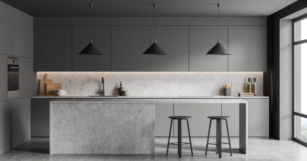 Een moderne, lichtgrijze keuken met minimalistische aankleding. De keuken beschikt over een grijze gietvloer, grijze, betonnen keukeneiland en warmgrijze keukenkasten. Drie donkergrijze hanglampen voegen een vleugje stijl en elegantie toe aan het sobere ontwerp.