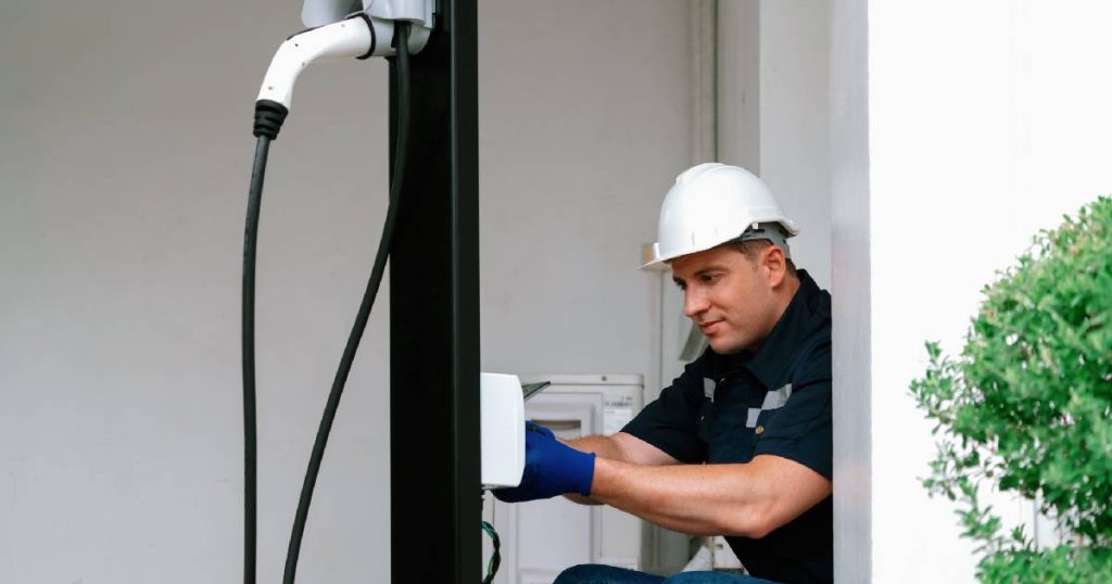 Een installateur sluit een loadbalancer laadpaal aan bij een familie thuis. Hij draagt een witte veiligheidshelm, blauwe handschoenen en een donkerblauw werkhemd.