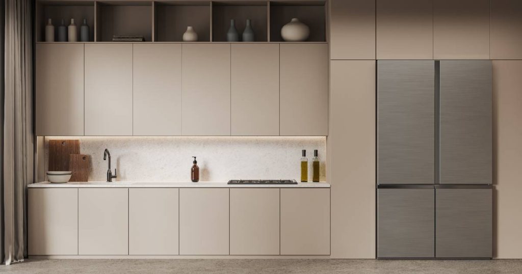 Een matte beige keuken met grote, opvallende grijs glanzende koelkast. De schappen boven de bovenkastjes zijn voorzien van decoratieve vaasjes in designkleuren: wit, licht beige en licht blauw.