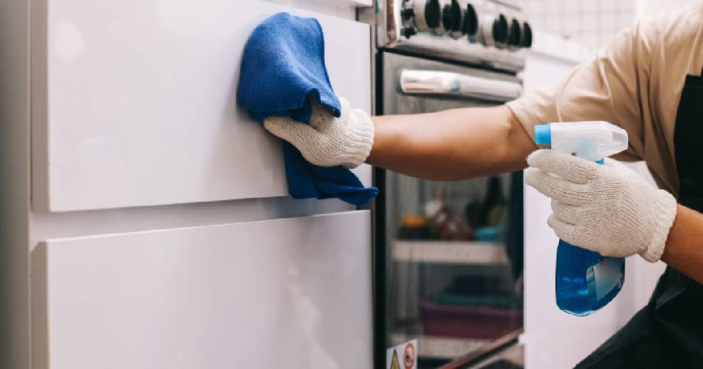 Een man met witte schoonmaakhandschoenen gebruikt een microvezeldoek, lauw water en een mild schoonmaakmiddel om de fronten van matte keukenkastjes schoon te maken en vette vingers te verwijderen.