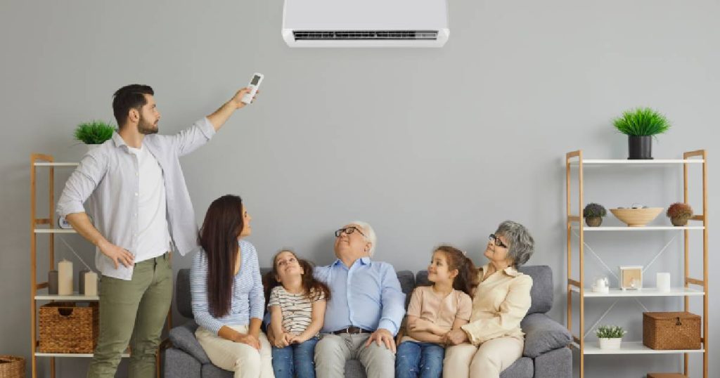 Een man (links) gebruikt een afstandsbediening om de binnenunit van een multi-split unit airco systeem te bedienen: een toestel dat je het hele jaar door controle geeft over het binnenklimaat in huis. Op de zetel onder de airconditioner unit zitten, van links naar rechts, een vrouw, een jong meisje, een oudere man, een jong meisje en een oudere dame.