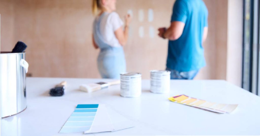 Een tafel met alle benodigdheden om een oude keuken te pimpen met verf: verfpotten, kleurstaaltjes en verfborstels. Op de achtergrond een dame en heer, beide gekleed in het blauw.