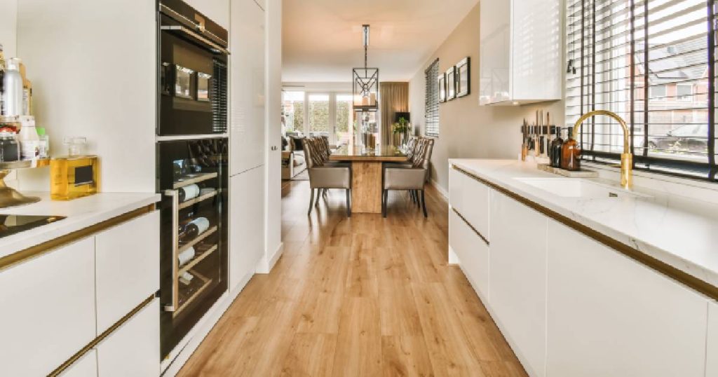 Een moderne witte parallel keuken met greeploze deuren, gouden kraan, raam en ingebouwde keukenapparatuur die uitgeeft op een eetkamer en woonkamer in een lange, smalle woning.