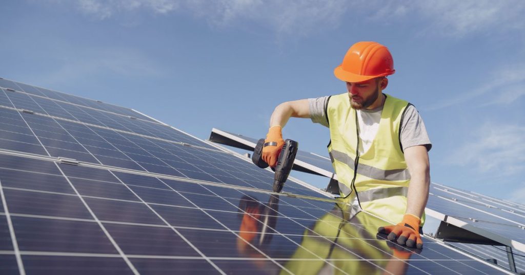 Man met oranje helm en geel hesje is met een boor zonnepanelen op een dak aan het schroeven