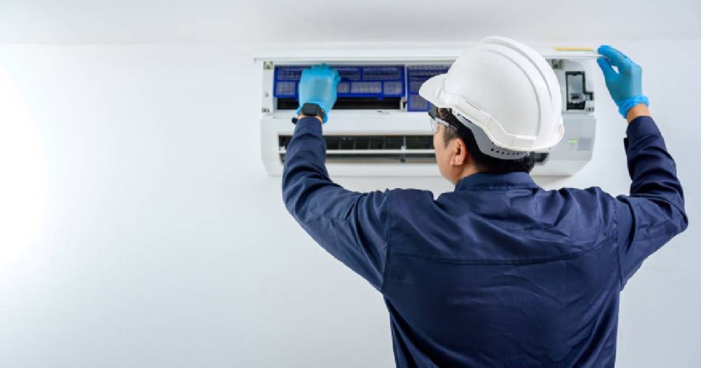 Een aircospecialist installeert de binnenunit van een airco met hoog rendement in een kamer met witte muren en een wit plafond. De specialist draagt een donkerblauw hemd, lichtblauwe werkhandschoenen en een witte helm met veiligheidsbril.