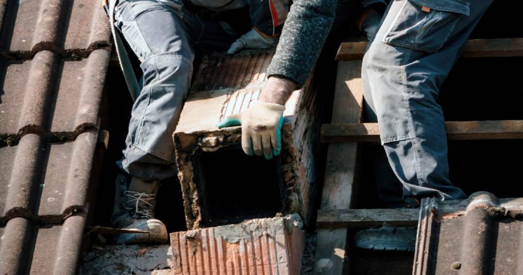 Twee vakspecialisten zijn in de weer met het afbreken en verwijderen van het bovendakse deel van een oude schouw.