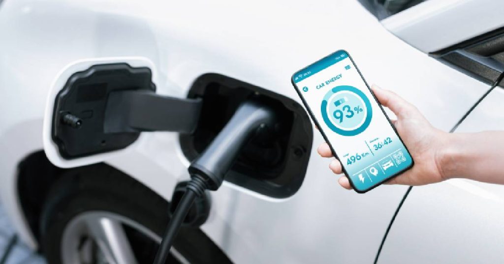 Een hand met smarthpone waarop de applicatie van een slimme laadpaal is geopend. Daarachter een witte elektrische auto met een zwarte laadkabel van diezelfde slimme laadpaal.