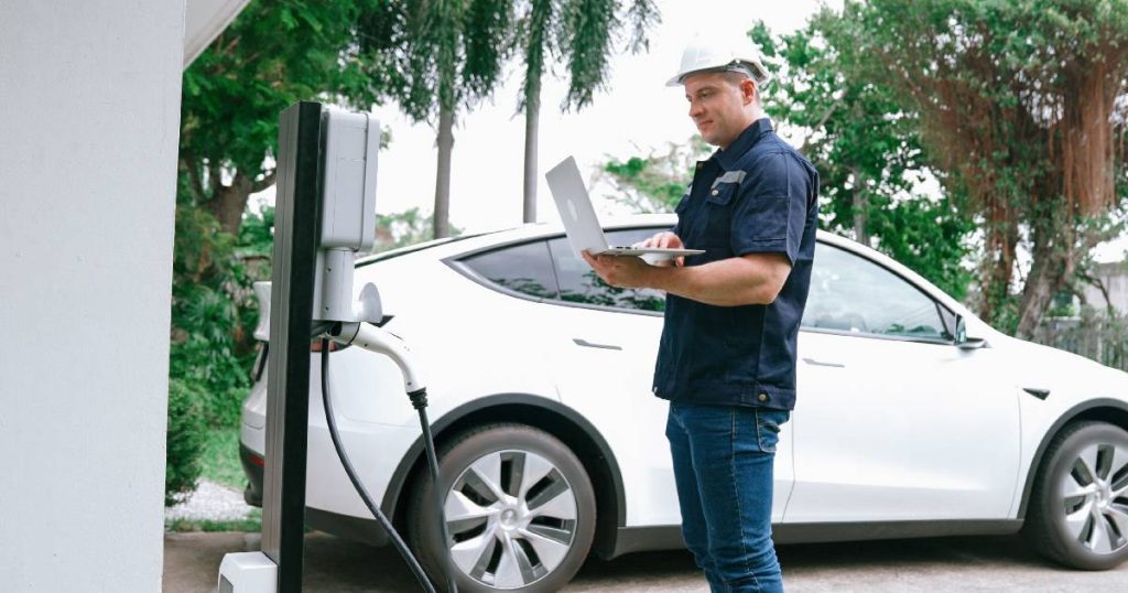 Een installateur installeert een snelle laadpaal bij een klant thuis. Op de achtergrond een witte elektrische auto en bomen.