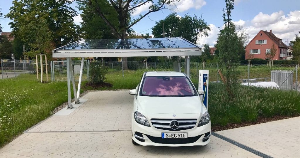 Een snelle laadpaal thuis, van elektriciteit voorzien door een zonnepaneelinstallatie op het dak van een carport. Naast de laadpaal staat een wit elektrisch voertuig.