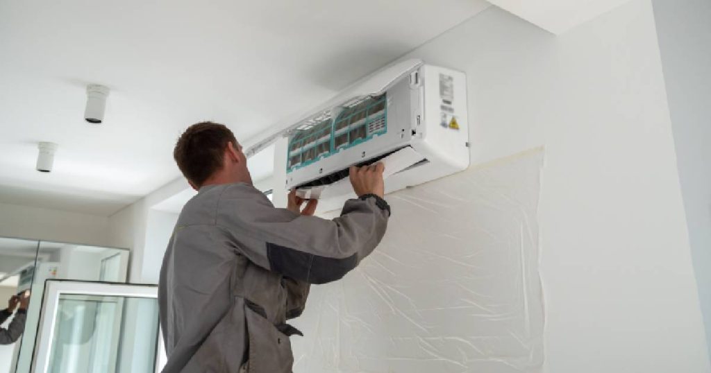 Een installateur in grijze werkkledij monteert de binnenunit van een split airco systeem. 