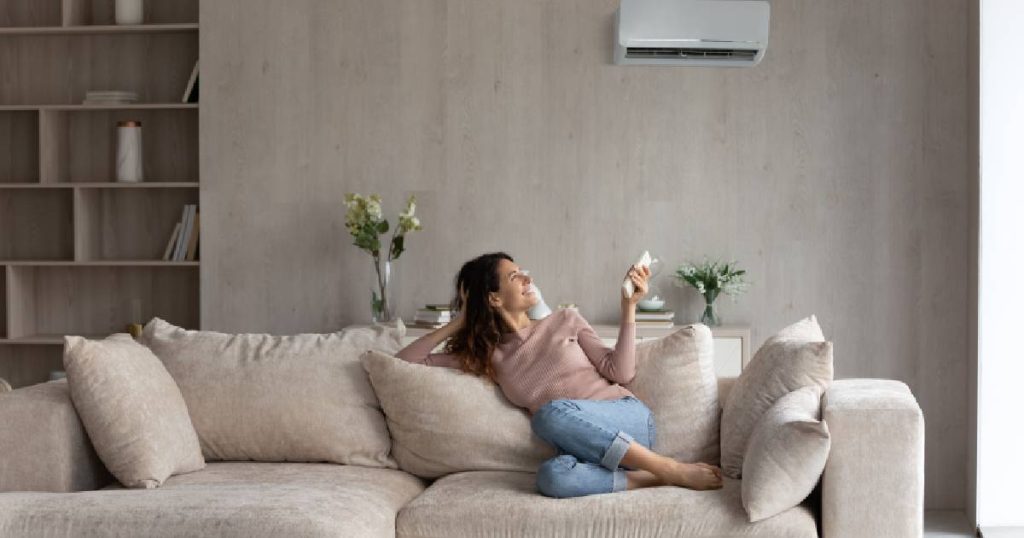 Een dame zit op een lichtbeige zetel in een beige living en gebruikt de afstandsbediening van haar airco om de temperatuur in huis aangenaam te houden tijdens de zomer.