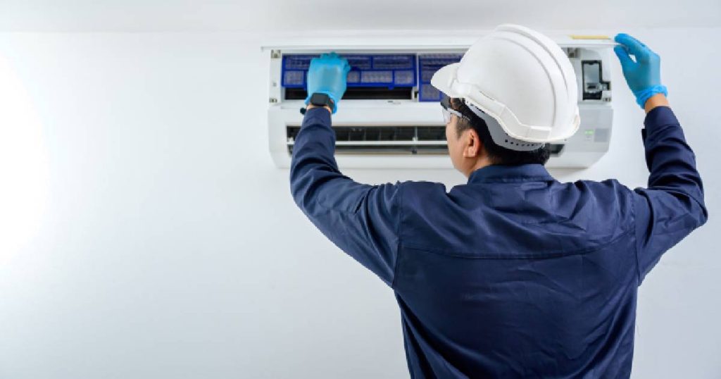 Een aircospecialist in blauwe overal en met een witte veiligheidshelm inspecteert de filters van een airco binnenunit. Dat airco onderhoud is cruciaal: een slecht onderhouden airco kan de temperatuur in huis niet efficiënt op peil houden.