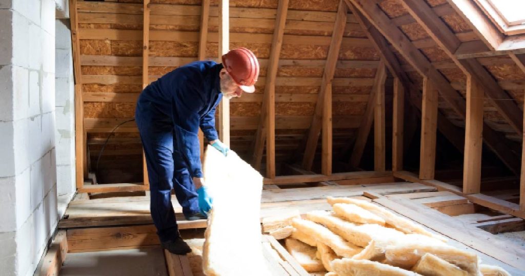 Een dakwerker is druk in de weer met het plaatsen van isolatiemateriaal in de zolderruimte onder een tentdak. Hij draagt een blauwe overall en oranjerode helm.