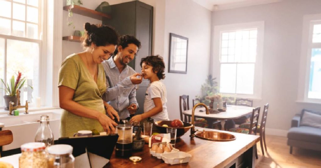 Een keuken zoals die van Dovy keukens, één van de top 10 keukenmerken, waar een vrouw, man en jong kind samen aan het koken zijn. De keuken beschikt over een houten keukeneiland met kleine koperen spoelbak en keukenkraan. De open keuken geeft uit op een eetruimte met living.