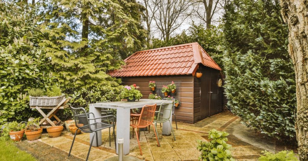 Tuinhuis met wolfsdak met oranje dakpannen. Tegen de gevel hangen bloempotten met rode bloemen.