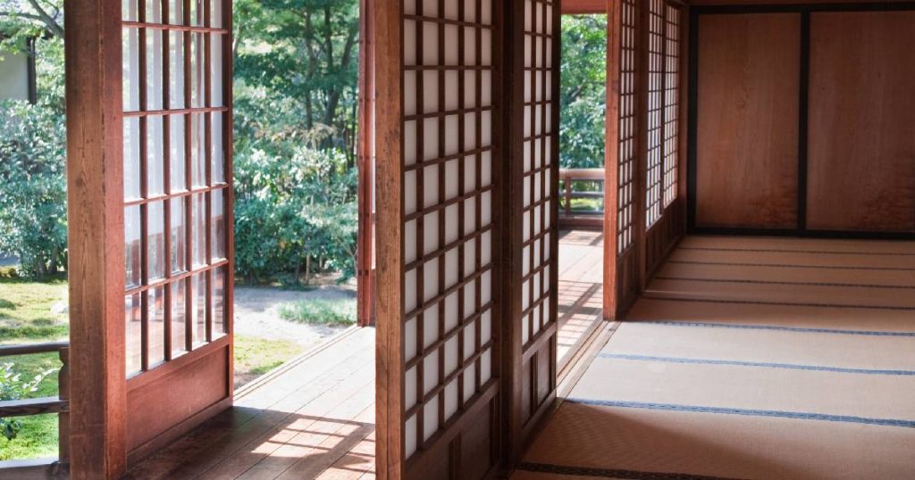 Schuin vooraanzicht van een ruimte die uitgeeft op een veranda. De veranda heeft men laten dichtmaken met hout en houten schuifdeuren met kleine raampjes in Japanse stijl. De veranda geeft uit op een groene tuin.