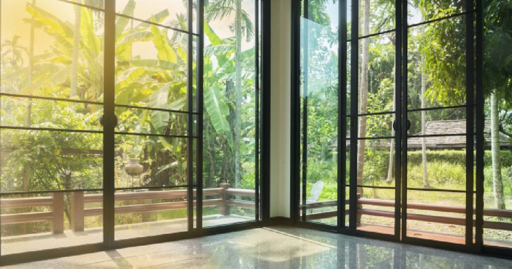 Een moderne veranda die men heeft laten dichtmaken met plexiglas. Het glas zit in een zwart, aluminium frame. De veranda geeft uit op een exotische, groene tuin.