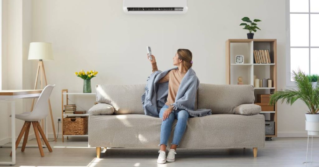 Een dame gebruikt de afstandsbediening om haar airco in te stellen op de gewenste temperatuur op een koude dag in de winter. Ze gebruikt een dekentje als extra verwarming: het is gezonder en energiezuiniger om het verschil tussen de temperatuur binnen en de temperatuur buiten niet te groot te maken met de airco.