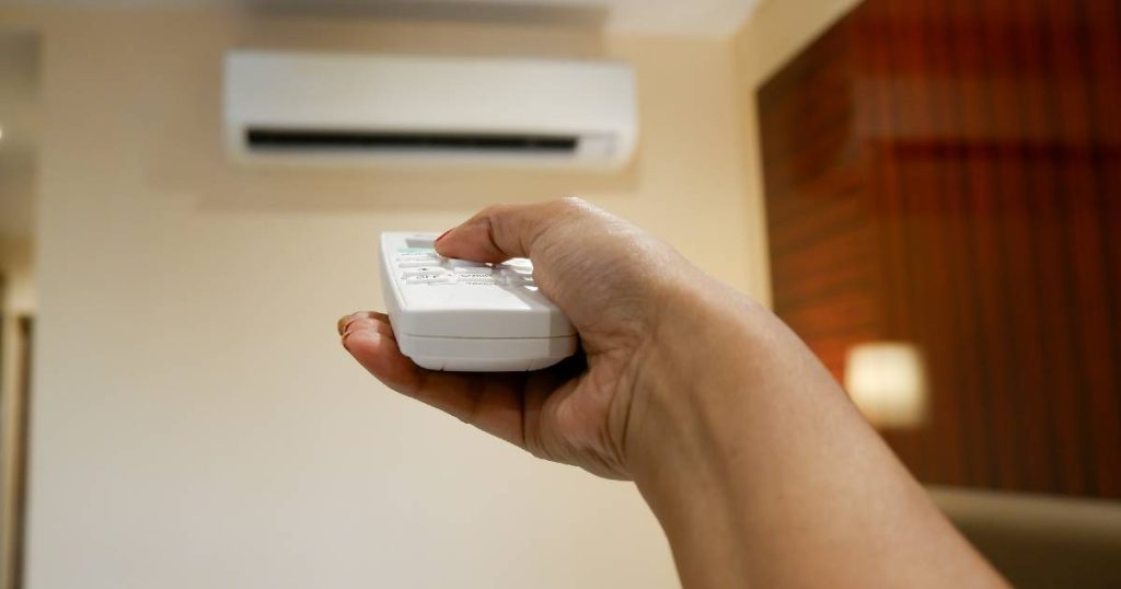 Een hand met afstandsbediening van een airconditioning binnenunit, met op de achtergrond het toestel zelf. 