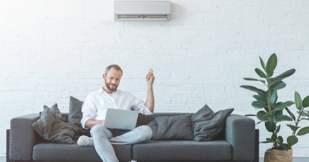 Een man zit met zijn laptop op de zetel en gebruikt de afstandsbediening om zijn airconditioner te bedienen. Hij beperkt het verschil tussen de temperatuur op de airco en de temperatuur buiten om kosten te besparen.