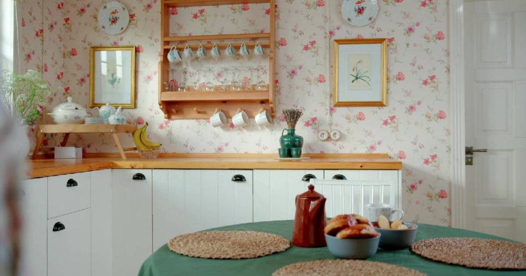 Een gezellige vintage keuken met landelijke uitstraling. Het behangpapier met bloemetjesmotief, het houten hangkastje, de ouderwetse schilderijtjes, de schattige witte keukenkastjes en het houten aanrecht nodigen uit tot gezellig tafelen.