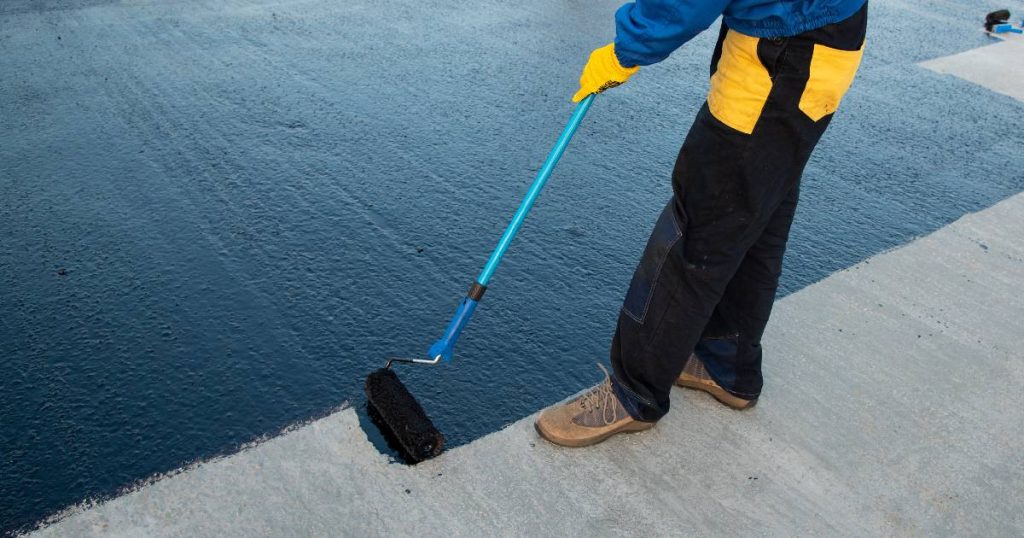 Een dakwerker brengt vloeibare bitumen dakbedekking aan op een betonnen dakvlak. Hij gebruikt een roller met telescoopsteel.