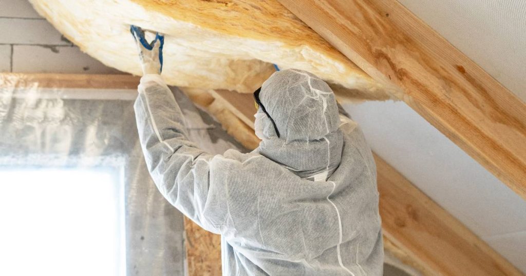 Een specialist in een wit beschermpak isoleert de binnenkant van een hellend dak om te voldoen aan de voorwaarden van de premie voor dakisolatie.