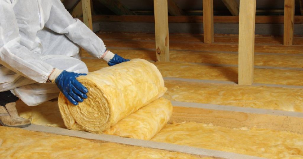 Een specialist in een wit pak met blauwe werkhandschoenen isoleert de zoldervloer om te voldoen aan de voorwaarden voor de premie voor dakisolatie.