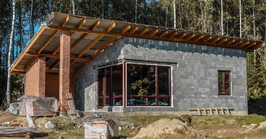 Een nieuwbouwwoning met lessenaarsdak onder constructie. De betonblokken en onderliggende dakconstructie zijn duidelijk zichtbaar, de grote ramen zijn alvast geplaatst.