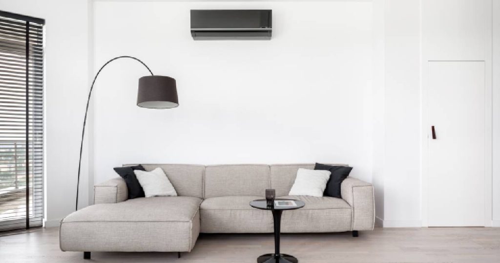 Een woonkamer met linksboven een Mitsubishi Electric airco in wandunit, een houten vloer, grijze zetels, een zwart bijzettafeltje en een zwarte designlamp. 