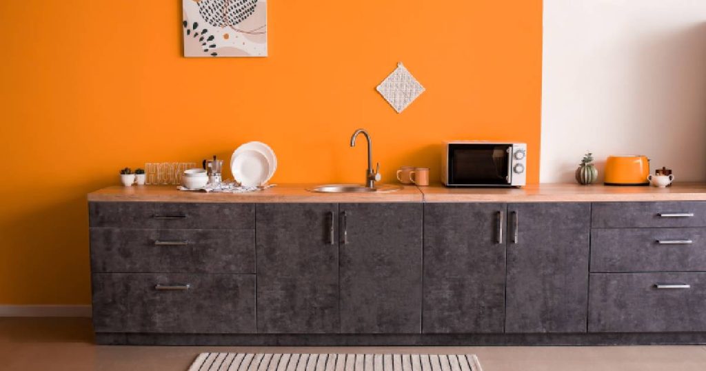 Een moderne keuken met donkergrijze of antraciet keukenkasten, houten aanrechtblad, beige vloer en opvallende oranje wand. Het oranje komt terug in de toaster en enkele kopjes. Een deel van de muur is wit: die accenten komen terug in de witte mini-oven en het servies alsook een wit keukenlapje en wit met oranje en beige schilderijtje tegen de oranje wand.