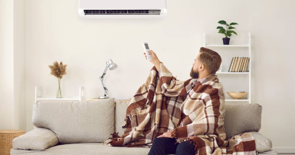 Een man in een dekentje gebruikt zijn airco om de temperatuur in huis comfortabel te houden tijdens de winter. Hij kiest met de afstandsbediening hoe hoog de temperatuur moet zijn.